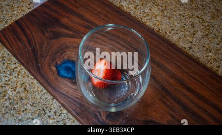 Strawberry sits inside a clear stemless wine glass resting on a dark wooden board Stock Photo