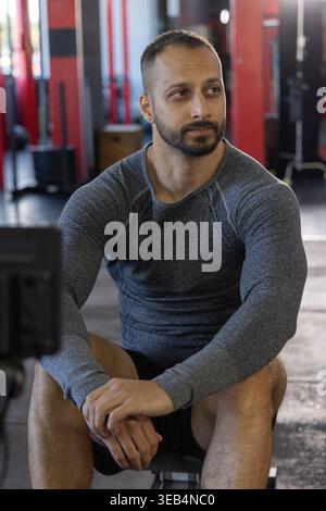 Indian man sitting and resting between sets on workout bench wearing grey top, black shorts. Gym, weights, fitness, equipment, athlete, focus, contemp Stock Photo