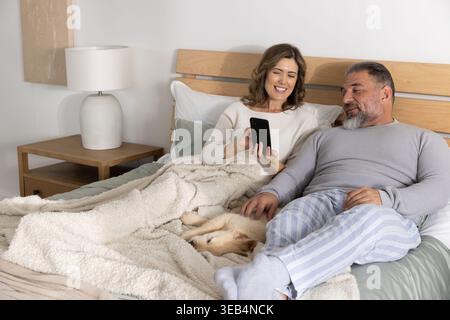 Couple with senior man and middle-aged woman reclining on bed, sharing phone and petting dog Stock Photo