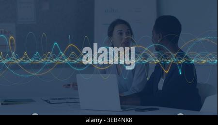 Discussing two colleagues in blazers gesturing over laptop at conference table, waveform overlay Stock Photo