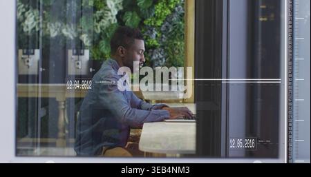 Working adult man typing on laptop at cafe window, wearing denim jacket, with plant wall overlays Stock Photo