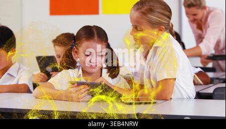 Using tablets, girls in white shirts leaning over desk in class with orange poster, yellow overlay Stock Photo