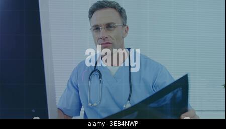 Inspecting doctor wearing blue scrubs holding X-ray and viewing monitor at clinic, with stethoscope Stock Photo