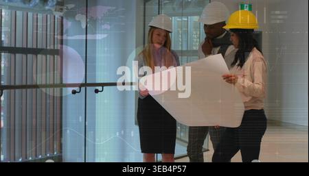 Studying three adults in hard hats holding large blueprint inside modern atrium with glass railing Stock Photo