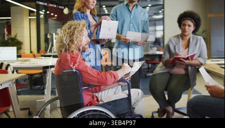Reading man in wheelchair in casual attire discussing printed page at office with overlaid charts Stock Photo
