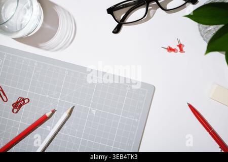Workspace with cutting mat, pencils, glasses, and office supplies on white desk. Copyspace Stock Photo