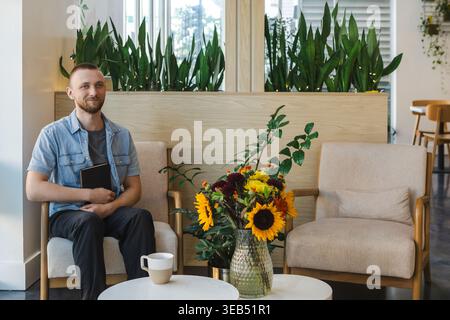 A man is seated in a room with plants around him. He has a notebook in his hands and is engaged in an online class. A vase with flowers and a cup are Stock Photo