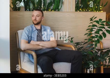 A young man with a thoughtful expression sits in a chair at a location filled with plants. He is engaged in remote work or online education. The atmos Stock Photo