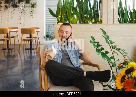 A man is seated in a cafe with plants in the background. He is talking on the phone and holding a drink. There are empty tables and chairs around him. Stock Photo