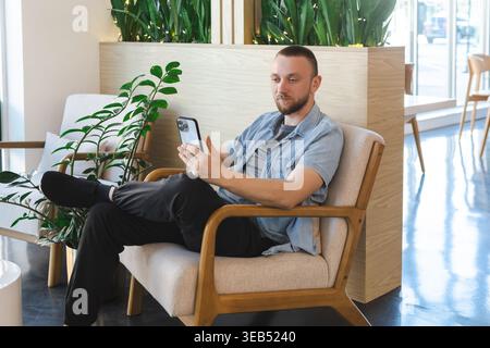 A man sits relaxed in a chair, holding a smartphone while focused on online lessons or a remote job task in a bright and modern interior space with pl Stock Photo