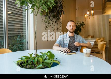A person is seated at a table in a modern workspace. They are focused on their notebook while a coffee cup rests nearby. Green plants add life to the Stock Photo