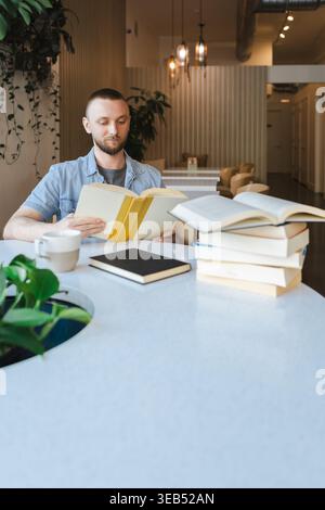 A person is sitting at a table in a bright workspace, reading books and working on an online course. Coffee and notebooks are on the table beside him. Stock Photo