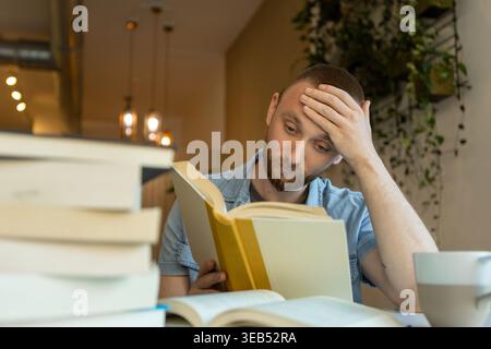 A man sits at a cafe with many books around him. He is focused on reading a book and appears to think deeply about what he is studying. It is daytime Stock Photo