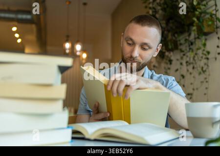 A man is reading a book at a table with stacks of books and a cup of coffee. He is focused on his study as he engages in online education. It is dayti Stock Photo