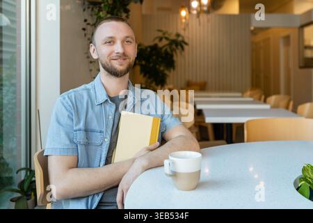 A man sits at a table in a coffee shop holding a notebook and a cup. He looks focused as he engages in online education or completes tasks remotely du Stock Photo
