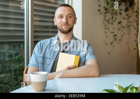 Person with short beard sits at a table with a cup of coffee. They hold a yellow notebook. Sunlight shines through a window. Green plants are nearby. Stock Photo