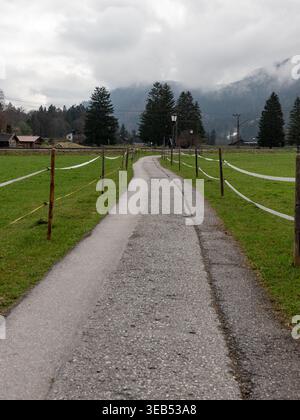 Wooden bridge and pathway leading toward alpine village across green meadow with mountain backdrop in Bavaria Germany. Stock Photo