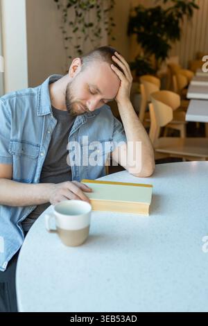 A person sits at a table with a book and a cup in a bright room. The individual appears focused and thoughtful while engaging in study or work. Stock Photo