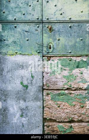 View of an old weathered wooden door with peeling green paint, metal rivets, and a galvanized metal patch in Italy. Stock Photo