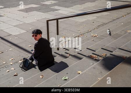 A young woman in sunglasses sits alone on sunlit urban steps with autumn leaves and a pigeon nearby in London, England Stock Photo