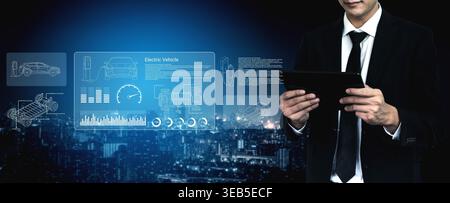 A businessman in a suit interacts with a tablet displaying electric vehicle data in a modern urban setting during the night, showcasing the Stock Photo