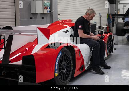 A mechanic in a black t-shirt sits on the sidepod of a red and white Le Mans prototype racing car while using a smartphone inside a professional pit g Stock Photo
