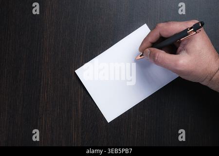 Hand holds pen, poised to write. White paper rests on dark wooden surface. Focused gesture suggests planning or creativity. Ideal for business, educat Stock Photo