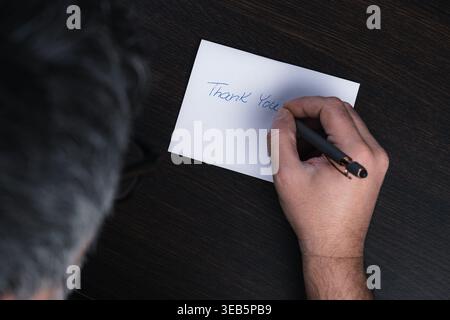 Hand writes "Thank You" on a white card. Dark table holds the card and pen. Person's hair and arm are visible from above. Simple gesture conveys grati Stock Photo