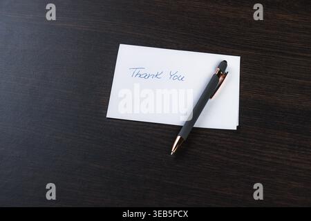 Simple thank you note rests on dark wood surface. Metallic pen lies beside the message, elegant and poised. Handwritten phrase shows appreciation, sin Stock Photo