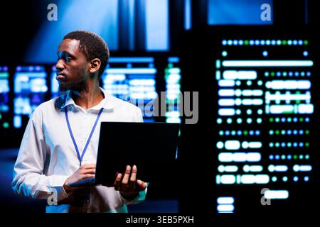 IT developer in server room using laptop to find firewall misconfigurations affecting mainframes performance and connectivity, leading to sluggish data transfer rates and poor client communication Stock Photo