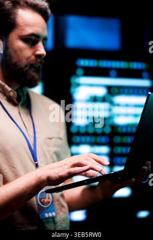 Serviceman doing system analysis, typing data on laptop of needed adjustments in supercomputers server farm housing advanced data storage infrastructure supporting critical IT workloads, close up Stock Photo