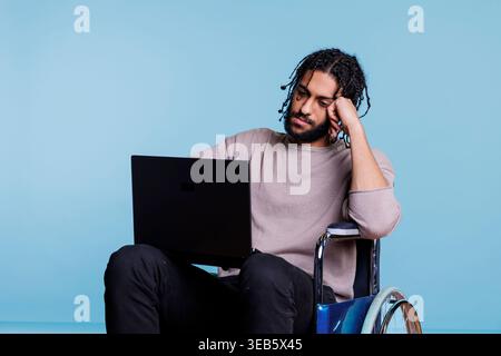Tired arab man in wheelchair typing on laptop while working remotely. Young exhausted freelancer software developer with physical imparment analyzing code on portable computer Stock Photo
