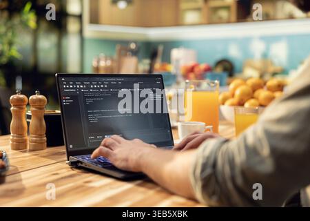 One person coding on laptop in a kitchen representing remote work in IT and software development. Programmer writes source code and uses command-line tools, highlighting engineering at home. Stock Photo