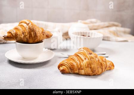 Freshly baked golden croissants, one balancing on a white coffee cup, and another resting on a light grey table, creating a delightful minimalist setu Stock Photo