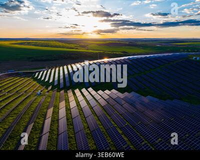 Solar panels cover a vast area of land under a bright sky. The sun sets and clouds float above the green hills. The scene shows renewable energy in ac Stock Photo