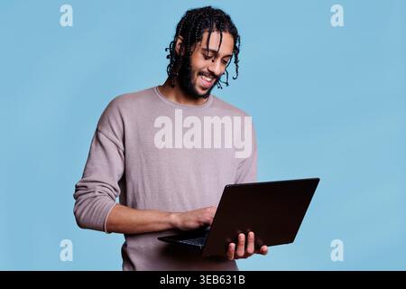 Happy arab man reading customer message and business report on laptop while smiling. Cheerful person wearing casual clothes holding portable computer and surfing internet Stock Photo