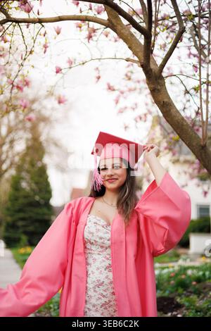 A young woman in a pink graduation gown poses joyfully while holding her cap. She stands in a garden with blooming trees, celebrating her achievement Stock Photo
