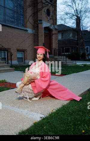 A graduate kneels on the pathway wearing a cap and gown. She holds a bouquet of flowers while celebrating her recent achievement. The setting is a sch Stock Photo