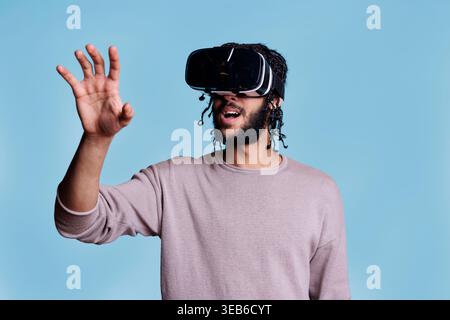 Man wearing vr headset exploring metaverse and making gestures with hands. Young person playing video game and enjoying cyber reality with virtual glasses on blue background Stock Photo