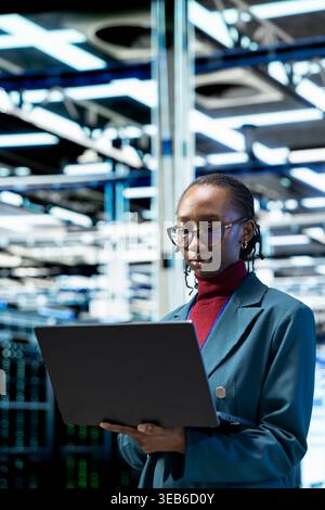 Data center female technician uses laptop to upgrade database storage equipment. African american woman in server room uses notebook to update cloud computing rigs systems, technology concept Stock Photo