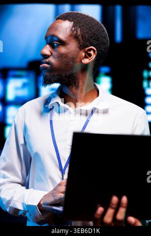 African american manager in server hub using laptop to find firewall misconfigurations affecting rackmounts performance and connectivity, leading to sluggish data transfer rates Stock Photo