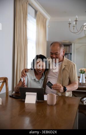 Senior Asian woman and Asian man leaning at dining table, using tablet, holding payment card Stock Photo
