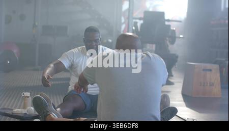 Stretching two African men in athletic T-shirts reaching forward on gym floor, with shaker bottle Stock Photo