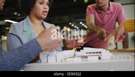 Collaborating team in gray blazer, pink polo, blue sweater pointing at scale model at studio table Stock Photo