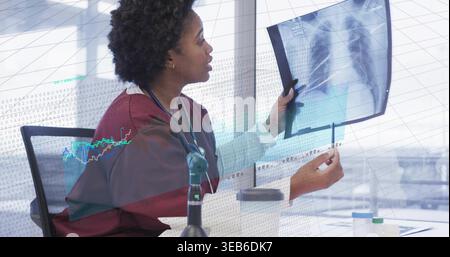 Examining woman in scrubs with stethoscope holding X-ray, pointing with pen at clinic desk, data Stock Photo