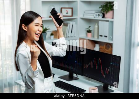Successful young Asian businesswoman earning high profit raising fist up, holding smartphone in exchange stock market's financial technology with Stock Photo