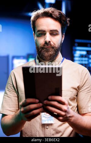 Vertical portrait of serviceman in server farm with supercomputers used for computationally intensive tasks such as scientific simulations. Employee doing annual equipment checkup using tablet Stock Photo