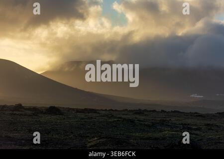 View of volcanic mountains partially covered by low clouds during golden hour over a rocky lava field in Lanzarote, Canarias, Spain. Stock Photo
