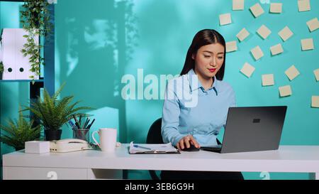 Smiling businesswoman typing data on laptop keyboard after cross-checking clipboard data notes. Focused professional employee working at modern creative office desk over blue studio background Stock Photo