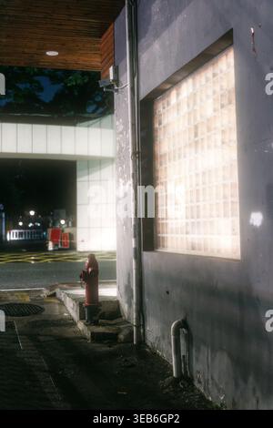 Nighttime urban alleyway with illuminated glass block window, red fire hydrant, concrete wall, and Stock Photo